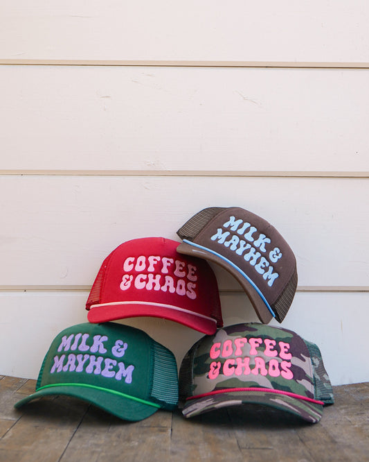 Four embroidered trucker hats reading "Coffee & Chaos" and "Milk & Mayhem" displayed on a rustic wooden table against a white wall.