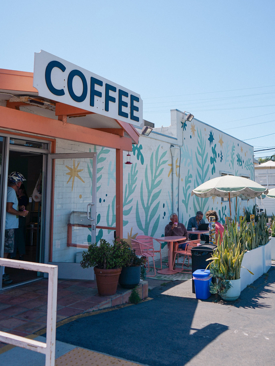 Colorful coffee shop exterior with a large “COFFEE” sign, mural-covered walls, outdoor tables, umbrella, plants, and customers.