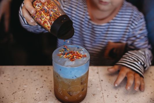 Child sprinkling colorful rainbow sprinkles onto a Birthday Cake Latte with blue cold foam at High Tide Coffee in San Clemente, celebrating the shop’s 7-year anniversary.