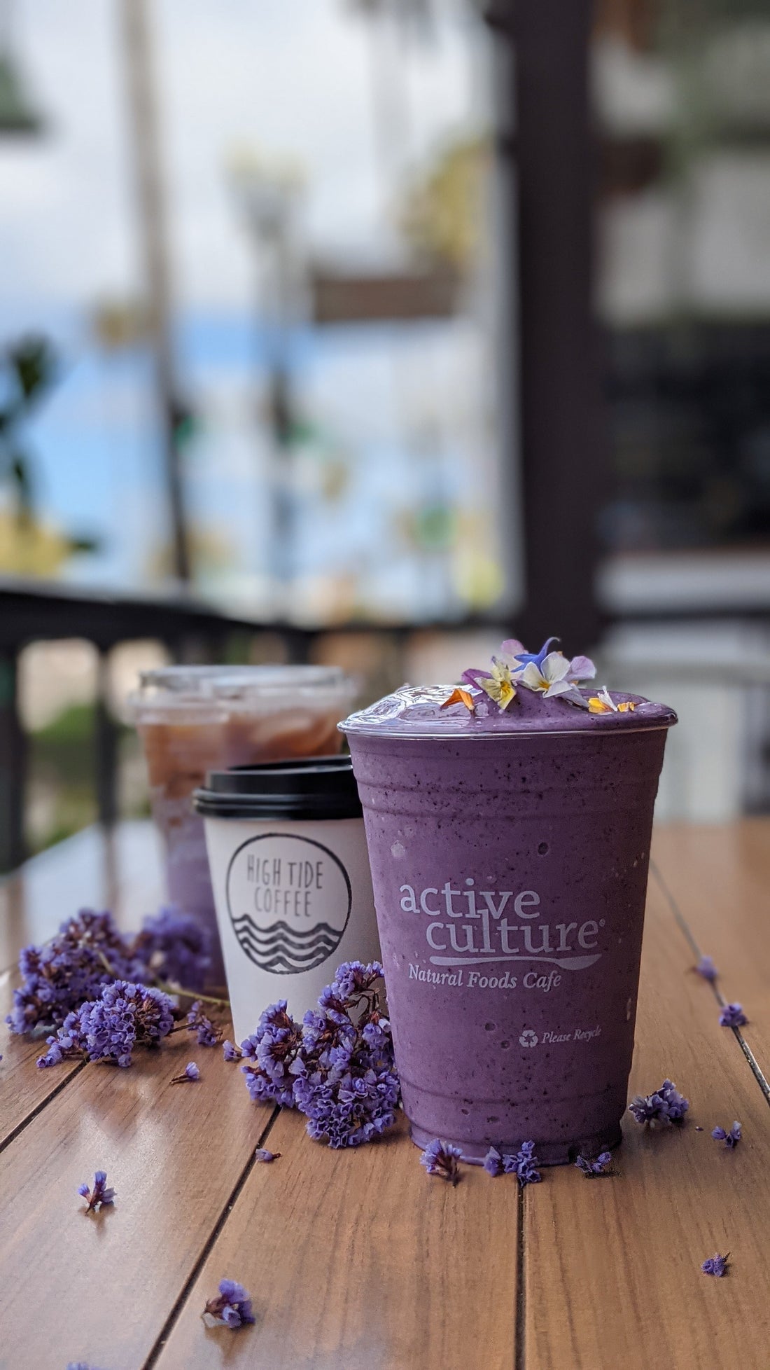 A vibrant purple smoothie topped with edible flowers sits on a wooden table next to a High Tide Coffee cup and iced coffee, surrounded by scattered purple blossoms.