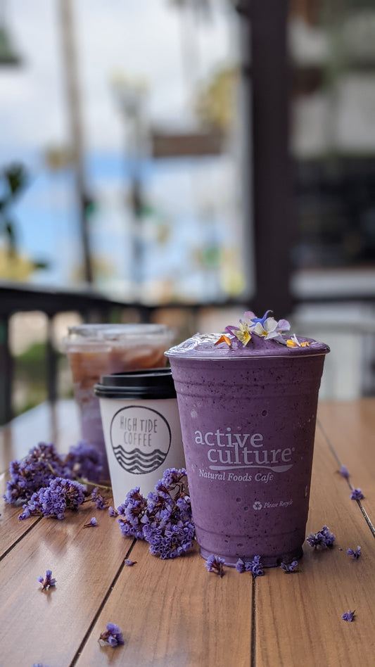 A vibrant purple smoothie topped with edible flowers sits on a wooden table next to a High Tide Coffee cup and iced coffee, surrounded by scattered purple blossoms.