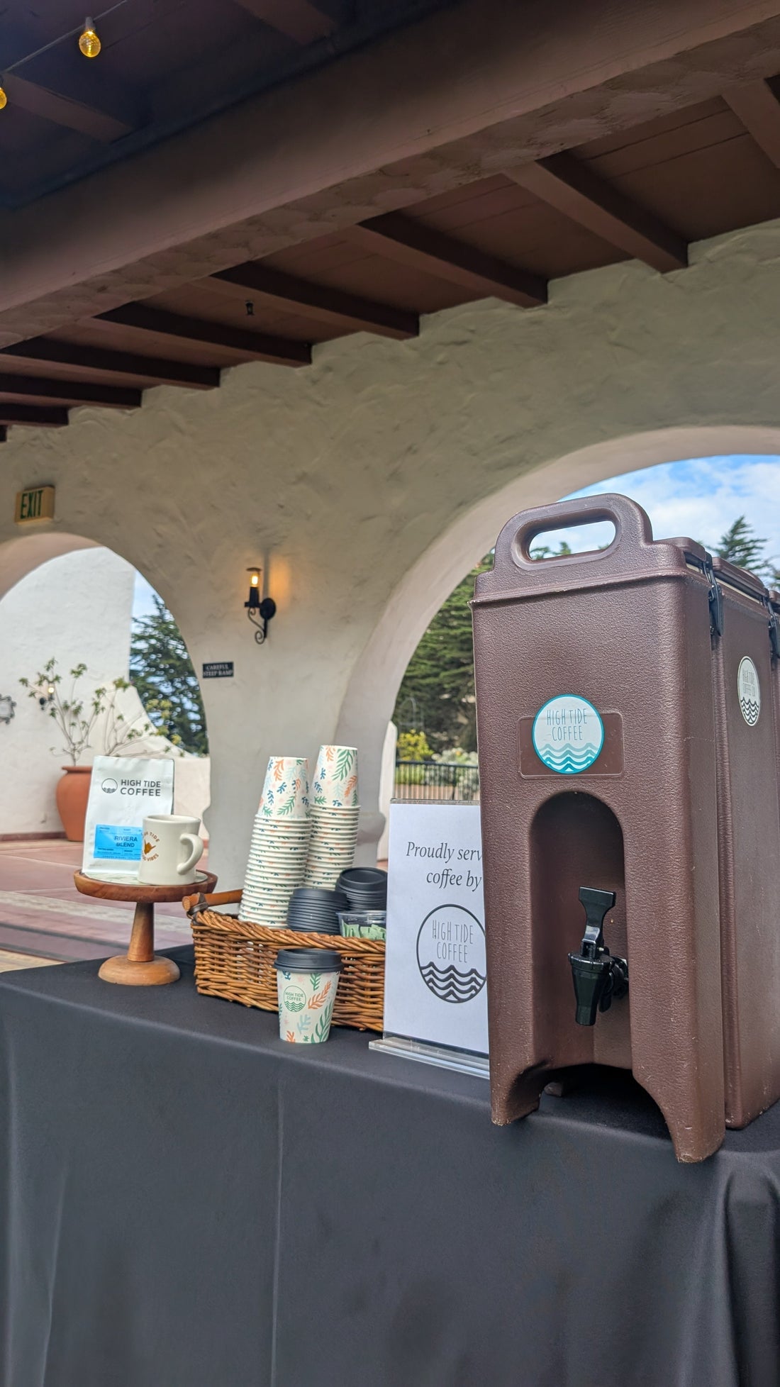 High Tide Coffee setup at Casa Romantica in San Clemente for the monthly Coffee Concerts, featuring a coffee dispenser, branded cups, a wicker basket of lids, and coastal-themed décor under Spanish-style arches.