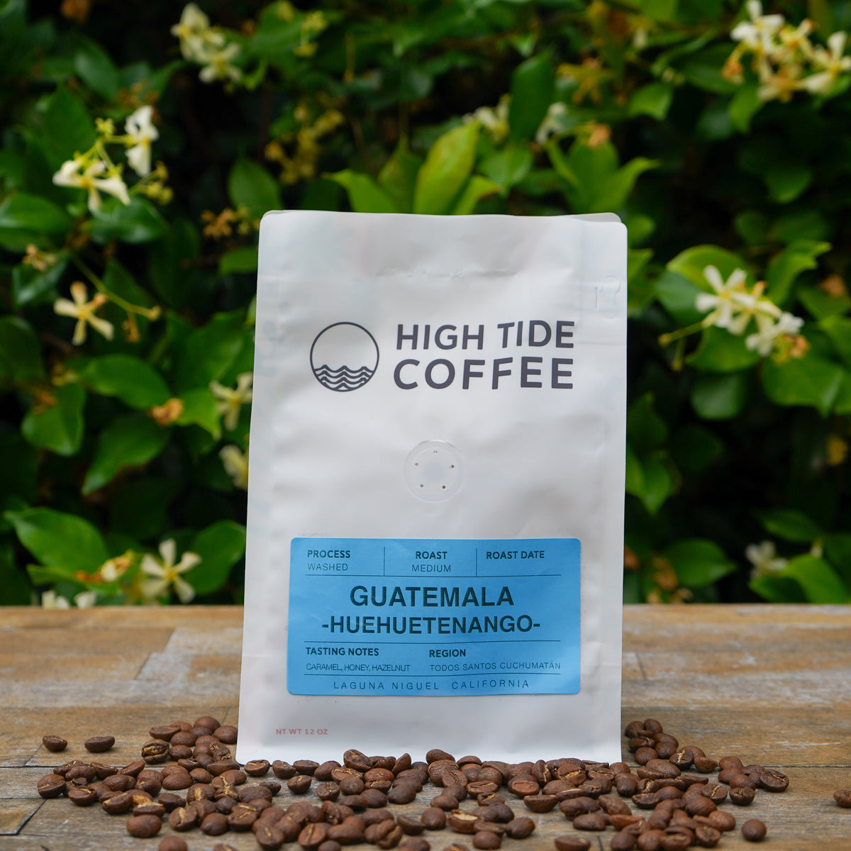 A bag of High Tide Coffee Guatemala Huehuetenango medium roast whole bean coffee stands on a wooden surface with scattered coffee beans in front, set against a background of green foliage and small white flowers.
