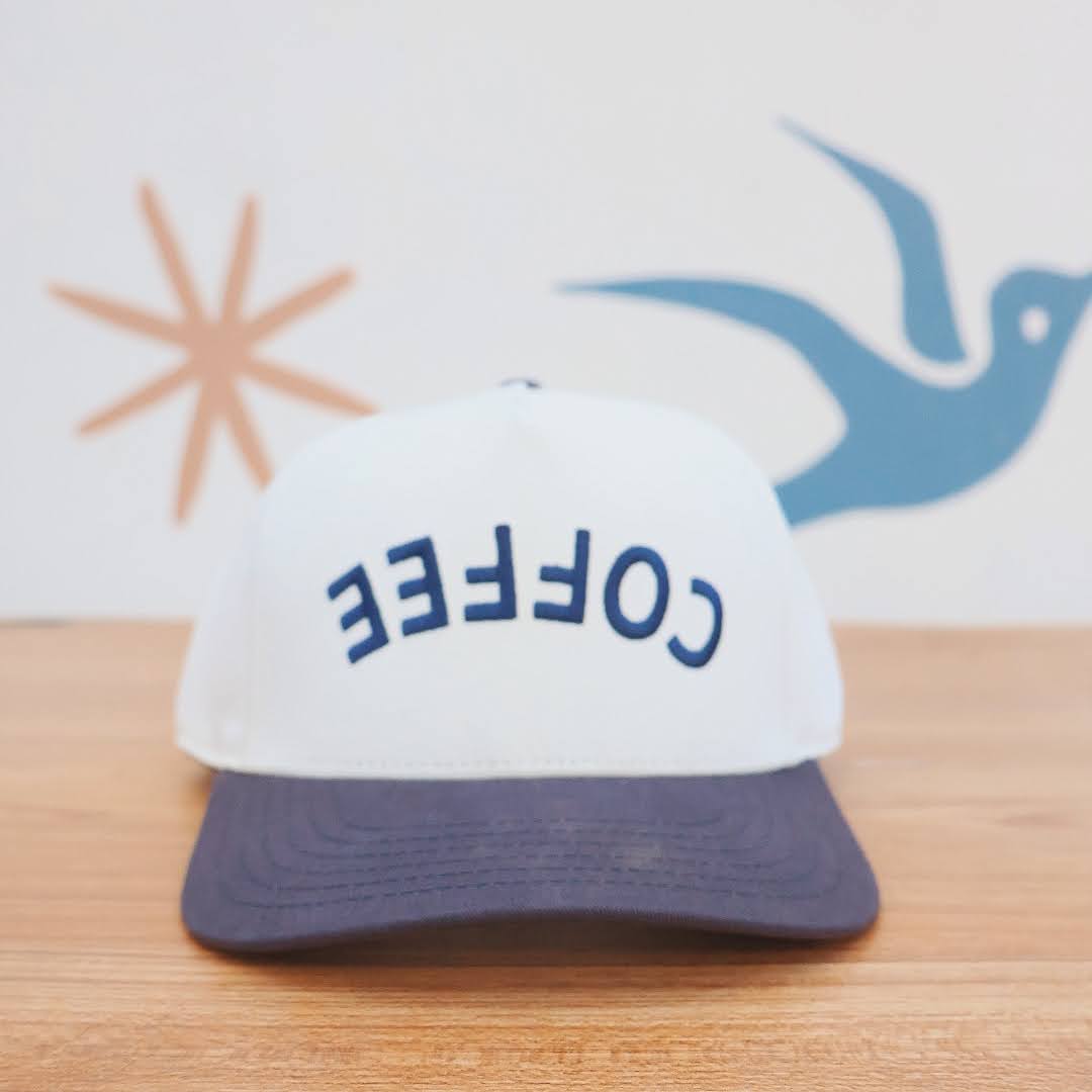 White cap with 'COFFEE' text on a wooden surface with abstract designs in the background
