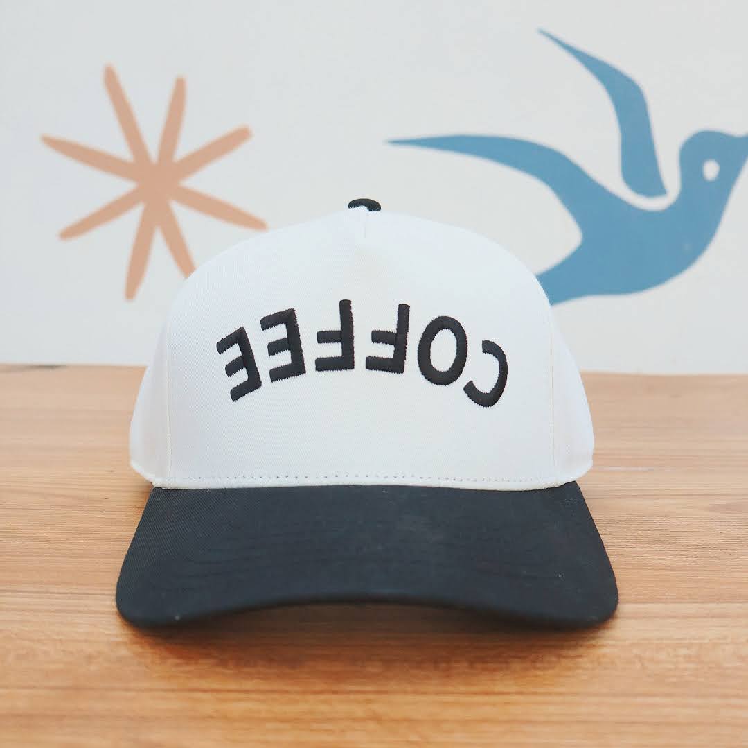 White cap with 'COFFEE' text on a wooden surface with abstract designs in the background