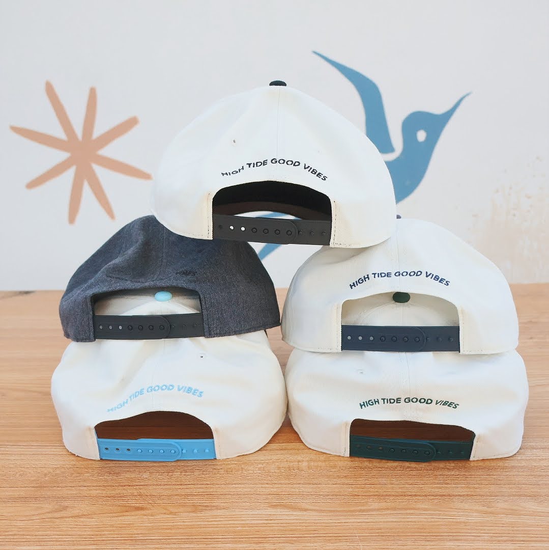 Three white caps with black and blue accents on a wooden surface, featuring 'High Tide Good Vibes' text.