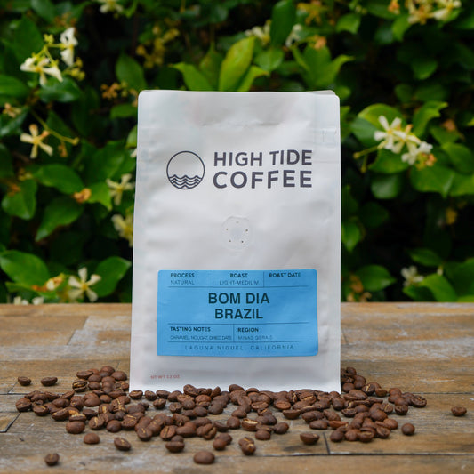 A bag of High Tide Coffee 'Bom Dia Brazil' whole bean coffee stands on a wooden surface with scattered coffee beans in front, set against a background of green foliage and small white flowers.