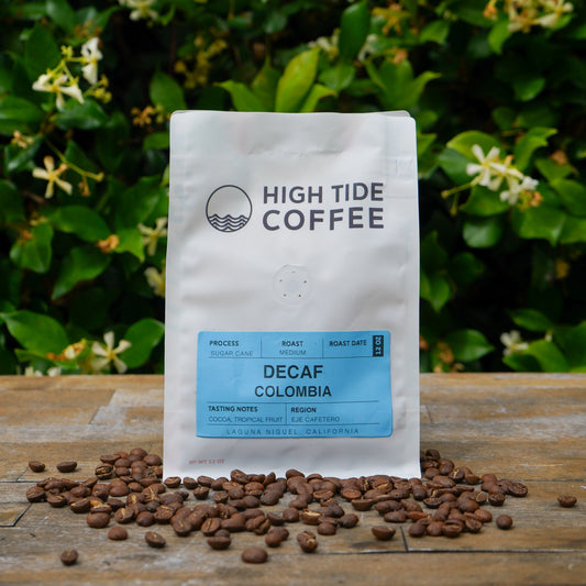 A white bag of High Tide Coffee Decaf Colombia coffee beans stands on a rustic wooden surface with scattered coffee beans in front, set against a background of green foliage and small white flowers.